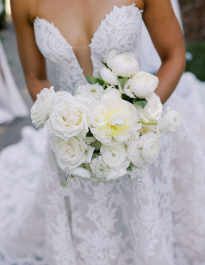 A bride in a strapless lace wedding gown holds a bouquet of white flowers, including roses and peonies.