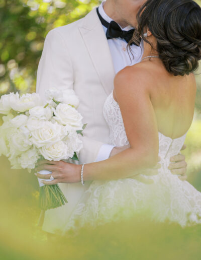 A bride in a strapless white gown holds a bouquet of white roses as she is embraced and kissed on the forehead by a groom in a white suit and black bow tie.