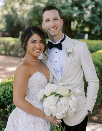 A bride in a white strapless dress and a groom in a white tuxedo jacket stand together outdoors, smiling and holding a bouquet of white flowers.