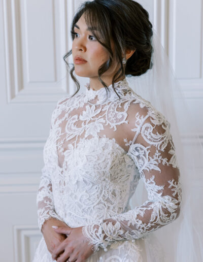 A woman in a long-sleeved, lace wedding gown with a high neckline and veil stands in front of a white paneled wall, looking off to the side.