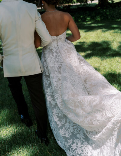 A couple is walking on grass, the person on the right is wearing a strapless white lace dress, and the person on the left is in a light-colored jacket and dark pants.