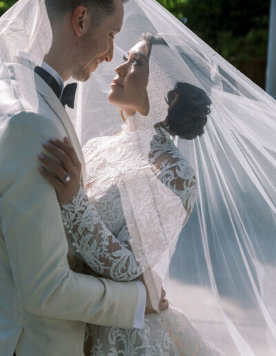 A bride and groom embrace under a lace veil outdoors, gazing at each other on their wedding day.