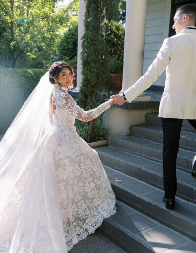 A bride in a lace wedding dress and veil holds hands with a groom in a white jacket as they walk up outdoor steps.