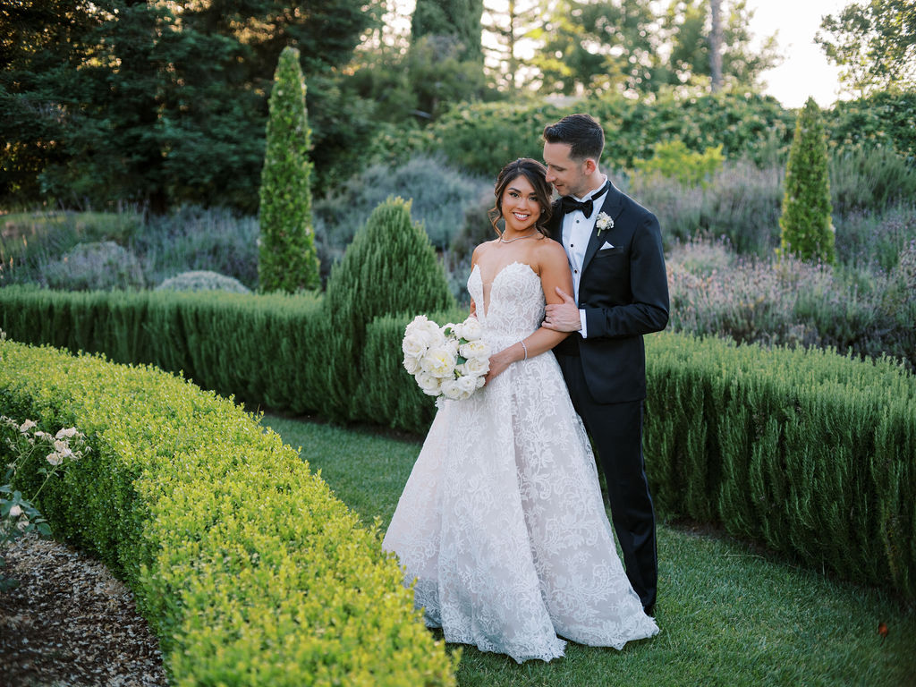 A bride in a white gown holds a bouquet while standing next to the groom in a tuxedo in a formal garden setting.