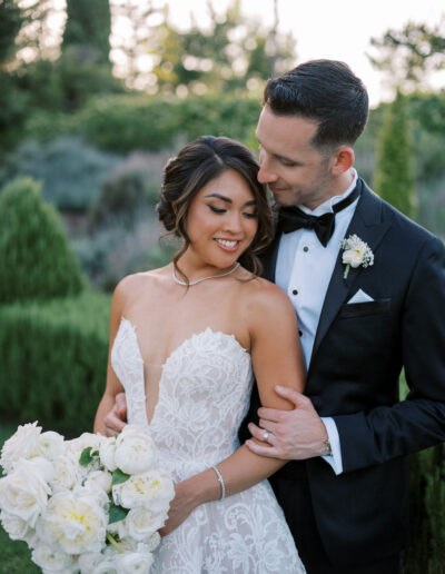 A bride in a lace wedding dress holding white roses stands with a groom in a black tuxedo, embracing outdoors in a garden.