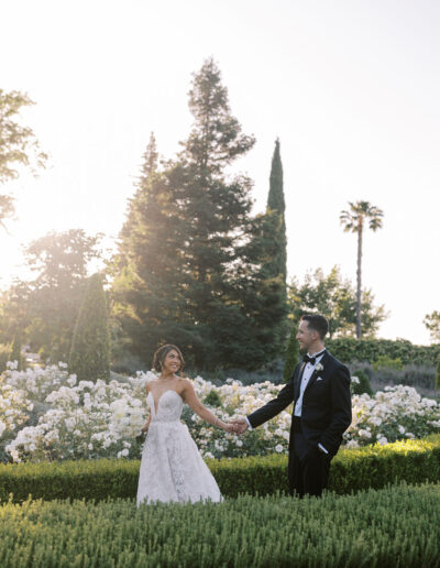 A bride and groom hold hands and face each other in a garden with blooming white flowers and tall trees in the background, dressed in wedding attire.