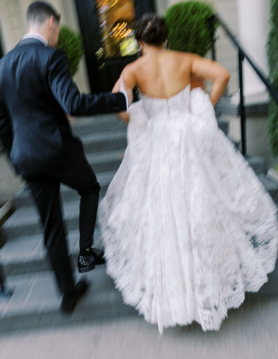 A bride in a white lace gown and a groom in a black suit walk up steps toward a building, holding hands. The image is slightly blurred, conveying movement.