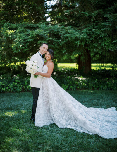 A bride and groom pose together outdoors, with the bride in a long lace gown holding a bouquet and the groom in a white jacket and black pants, standing on grass in front of greenery.