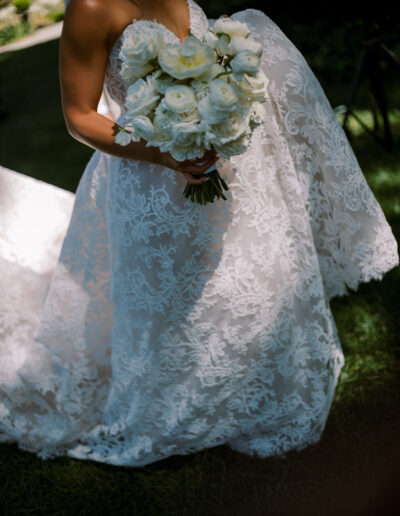 A person in a white lace wedding dress holds a bouquet of white flowers outdoors.