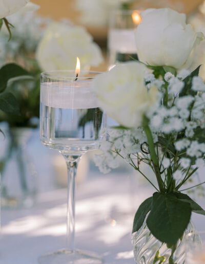 A floating candle in a glass of water is surrounded by white roses and baby's breath flowers in small glass vases on a white tablecloth.