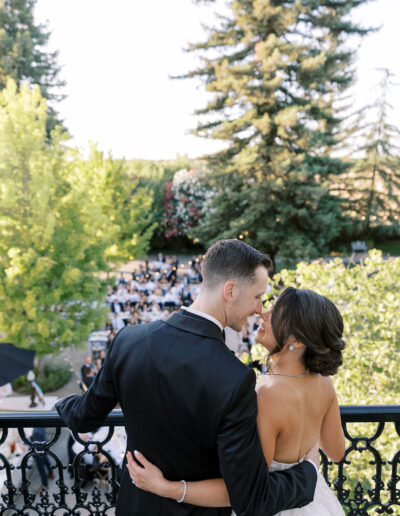 A couple in formal attire stands on a balcony, embracing and looking at each other, with a wedding reception and trees visible in the background.