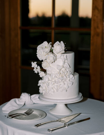 A two-tier white cake decorated with white flowers sits on a cake stand on a round table with a napkin, fork, knife, and cake server.
