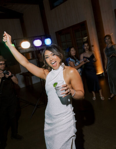 A woman in a white dress holds a drink and raises her arm while smiling at a party, with people and colorful lights in the background.