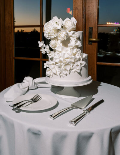 A white two-tiered cake decorated with white flowers sits on a round table set with a napkin, plate, fork, knife, and cake server in front of glass doors at sunset.