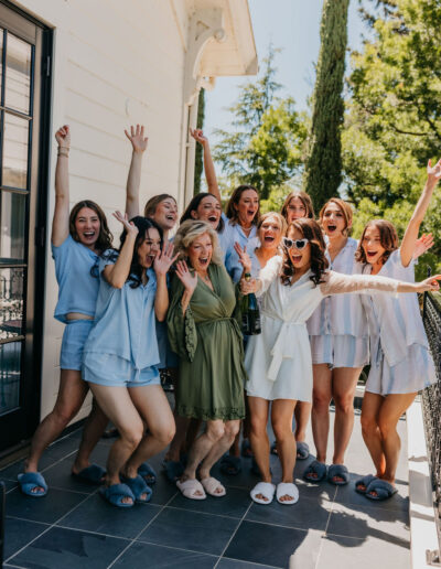 A group of women, some in matching light blue pajamas and slippers, stand on a patio cheering and smiling around a woman in a white robe and a woman in a green robe.
