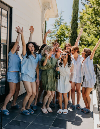 A group of women in sleepwear and slippers stand on a patio, smiling and celebrating with raised hands and a bottle of champagne.
