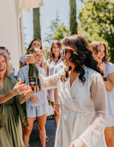 A woman in a white robe and sunglasses holds up a bottle of bubbly, surrounded by smiling women outdoors on a sunny day.