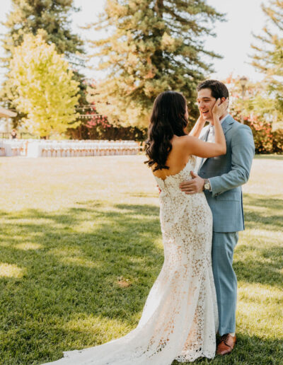 A bride in a lace dress and a groom in a light blue suit embrace and smile at each other outdoors on a sunny day, with trees and grass in the background.
