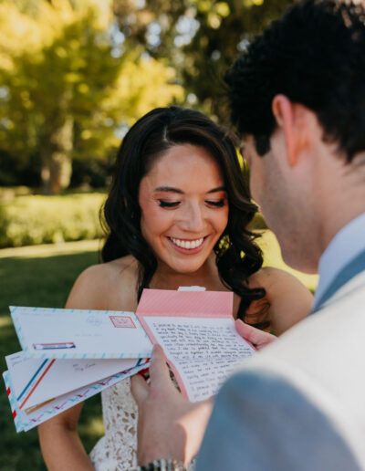 A woman in a white dress smiles while a man in a suit reads a handwritten letter to her outdoors, holding several colorful envelopes.