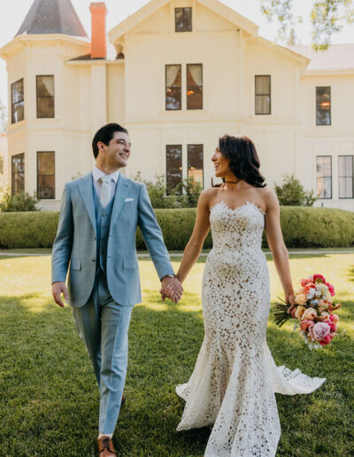 A bride in a white lace dress and a groom in a light blue suit walk hand in hand on grass in front of a large, light-colored house.