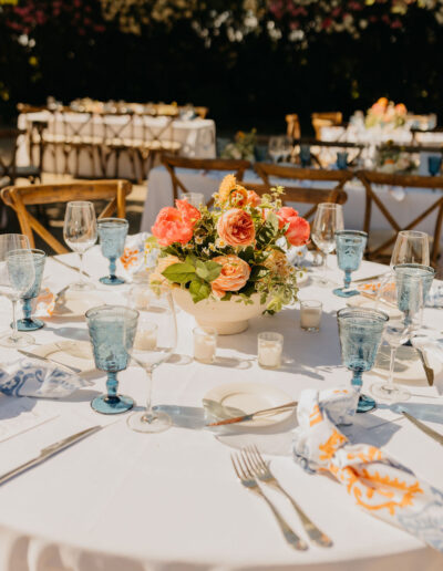 A round table set for an outdoor event features blue glasses, white plates, silver cutlery, and a floral centerpiece with orange and yellow flowers.