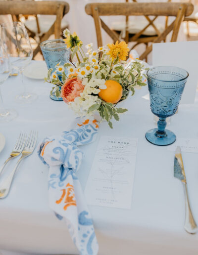 A table setting with blue glassware, gold cutlery, a floral centerpiece with citrus fruits, a printed menu, and a cloth napkin with blue and orange patterns.