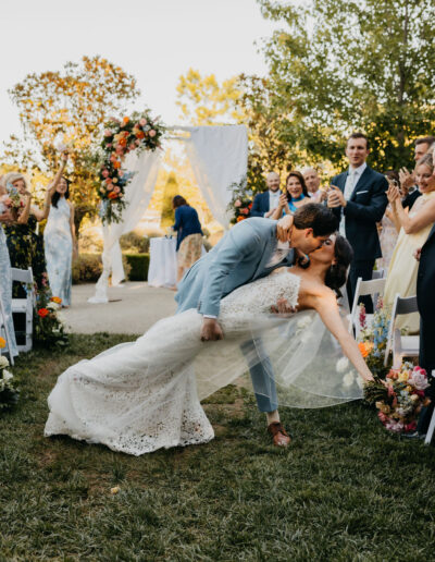 A groom in a blue suit dips and kisses the bride in a white lace gown during an outdoor wedding ceremony, with guests applauding and floral decorations in the background.