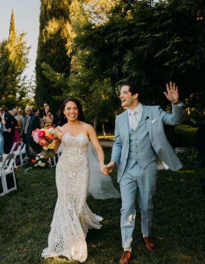 A bride in a white lace gown and a groom in a light blue suit walk hand in hand outdoors, smiling and waving, surrounded by wedding guests.