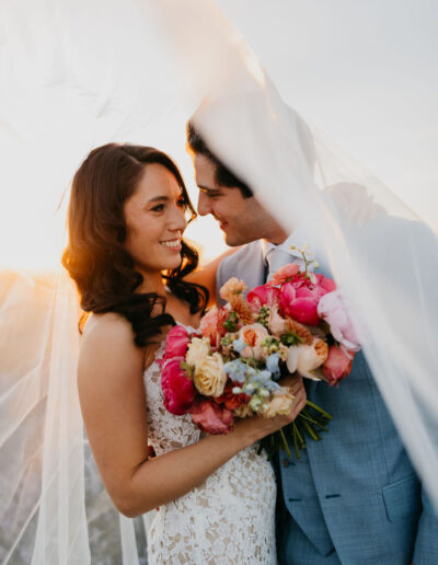 A bride in a lace dress and groom in a light blue suit stand close together, holding a colorful bouquet under a flowing veil at sunset.