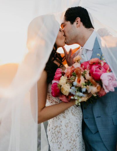 A bride and groom kiss under a sheer veil; the bride holds a colorful bouquet of flowers, and the groom wears a light blue suit.
