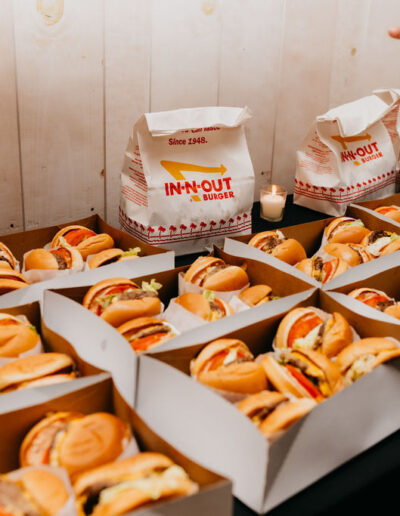 Multiple open boxes filled with In-N-Out burgers are arranged on a table; a hand points toward the food, and candles are visible in the background.
