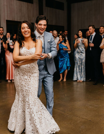 A newlywed couple, dressed in a white lace gown and light suit, dance together in the center of a room while a group of guests stand around watching and smiling.