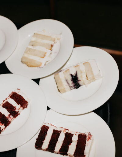 Several white plates with slices of layered cake, including red velvet and white cakes, arranged on a dark surface.