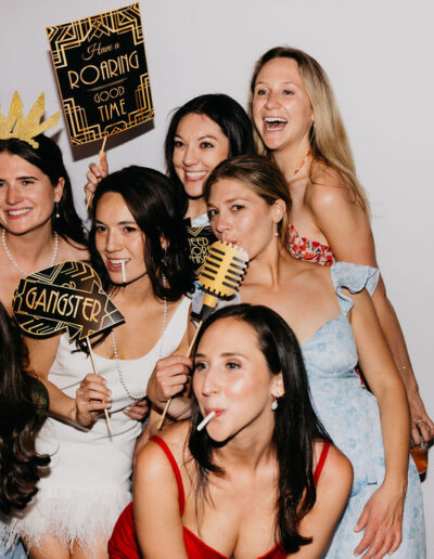 Six women pose together in a photo booth, holding props and signs, including "Have a Roaring Good Time" and "Gangster," with playful expressions and lollipops.