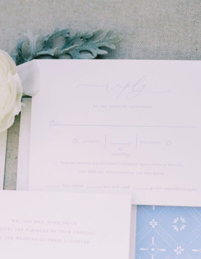 A flat lay of wedding stationery, including an RSVP card, an invitation, a blue patterned card, and a white ranunculus flower on a light fabric background.