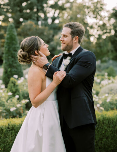 A bride and groom stand outside in a garden, gazing at each other and embracing, dressed in a white gown and black tuxedo.