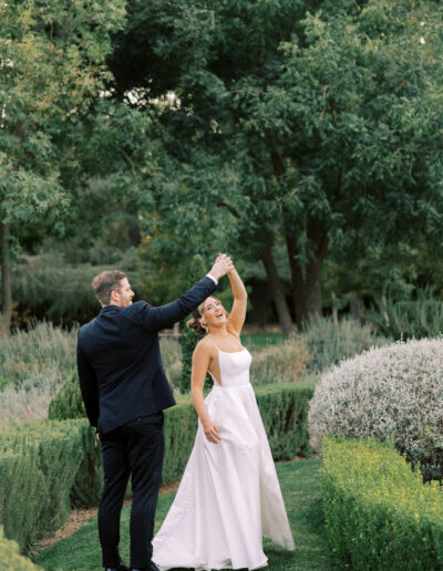 A bride in a white dress and a groom in a black suit dance together outdoors on a green lawn surrounded by bushes and trees.