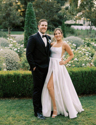 A man in a black tuxedo and a woman in a white gown pose together outdoors in a manicured garden with green hedges and flowering bushes.