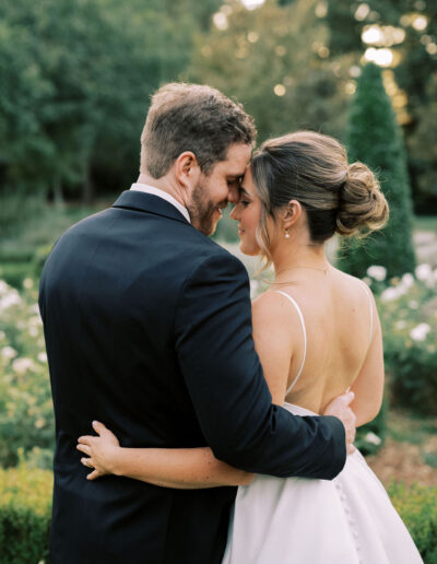 A bride and groom embrace outdoors, standing close with their foreheads touching, surrounded by greenery and white flowers.