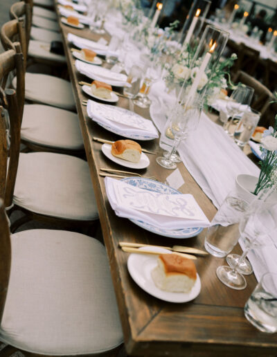 A long wooden table is set for a formal meal with plates, bread rolls, glasses, candles, and floral centerpieces.