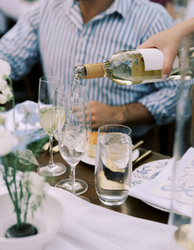 A person pours white wine into a glass at a table set with water, wine glasses, white flowers, and a man in a striped shirt.