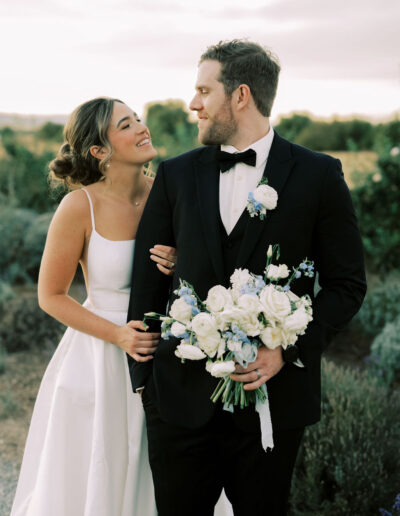 A bride and groom stand outdoors, the bride smiling at the groom who is holding a bouquet of white and blue flowers. Both are dressed in formal wedding attire.