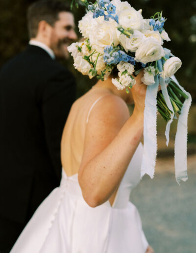 A bride in a white dress holds a bouquet of white and blue flowers, while a man in a black suit stands beside her, both outdoors.