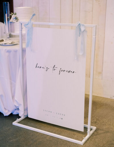 A white wedding sign with blue ribbon reads "here's to forever" and displays the names "Jacob + Layne" and the date "10 . 14 . 23" in a modern, minimal font.