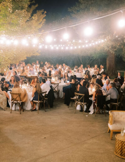 Outdoor evening wedding reception with guests seated at tables under string lights, children in white dresses, and decorated tables and lounge area on a patio surrounded by trees.