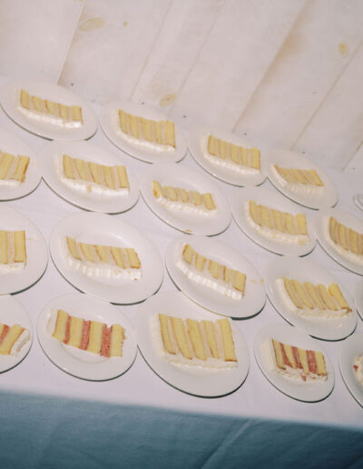 Several plates with slices of layered cake are arranged in rows on a white tablecloth next to a tall candleholder and a metal container.