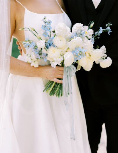 A bride in a white dress holds a bouquet of white and light blue flowers, standing next to a person in a black suit.