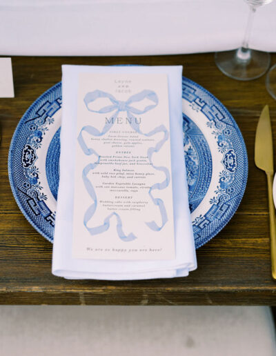 A formal table setting with a blue-patterned plate, gold utensils, a white napkin, a printed menu, wine glasses, and name cards on a wooden table.