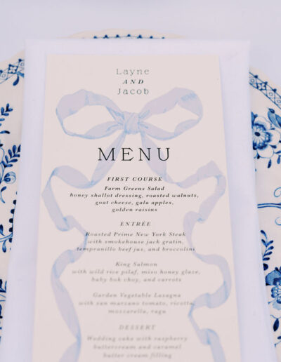 A printed wedding menu with blue ribbon design is placed on a white plate with blue floral patterns, accompanied by gold cutlery.