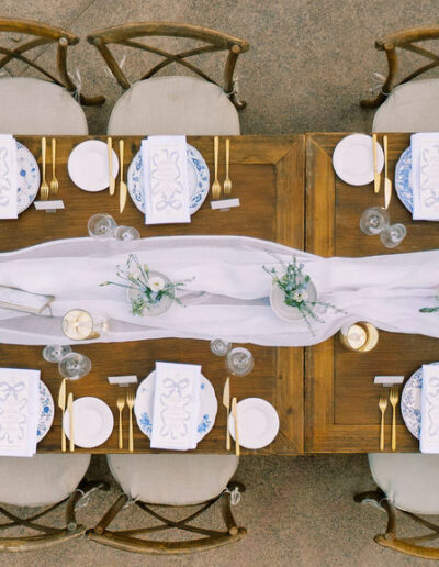 Aerial view of a long wooden dining table set for an event with white plates, blue napkins, gold cutlery, floral centerpieces, and a white table runner.